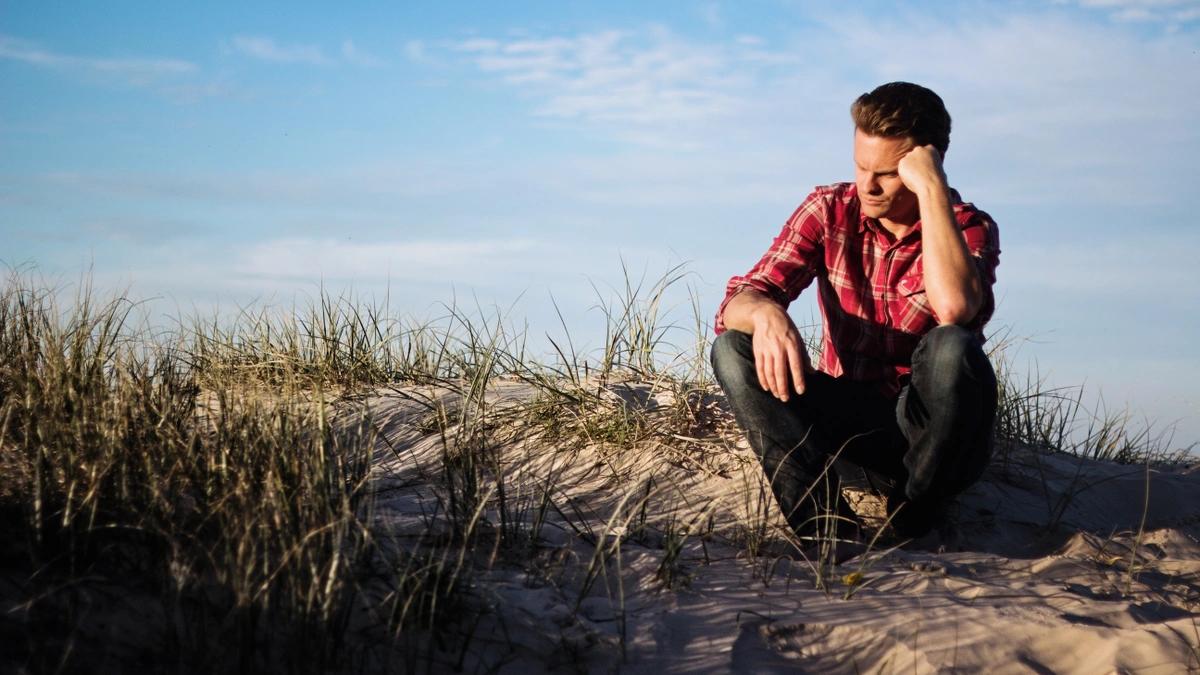 Man in red flannel shirt and jeans sits on a grassy sand dune with his head on his hand, appearing discouraged.