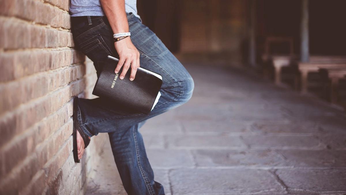 Man leans back against a brick wall while standing and holding a bible in his right hand. The picture is from the side and cropped to only show his waist down.