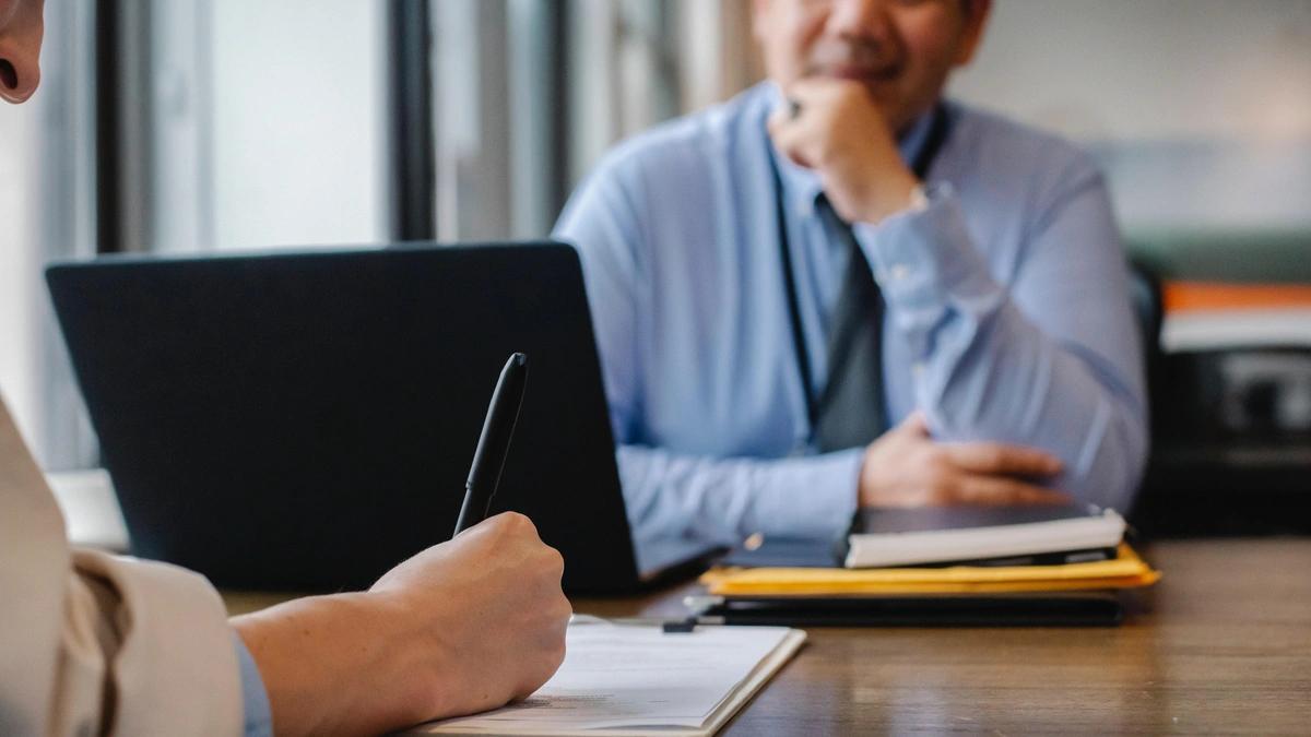 zoomed in image of two people at a table with notebooks and a laptop, one person taking notes while the other looks up with hand on chin