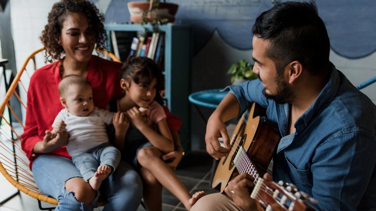 Dad plays guitar while mom sits across from him holding their two kids.